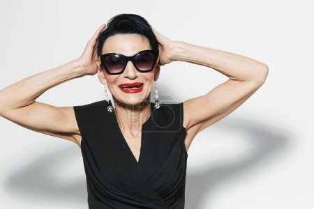 Stilvolle ältere Frau in Sonnenbrille und schwarzem Kleid posiert selbstbewusst mit den Händen auf dem Kopf für ein Porträt vor der Kamera