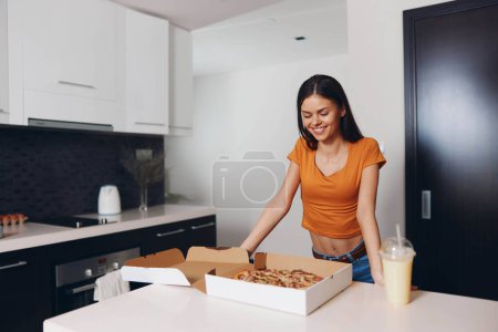 Téléchargez les photos : Femme debout près de la boîte à pizza ouverte avec boisson sur le comptoir dans l'intérieur de la cuisine confortable - en image libre de droit