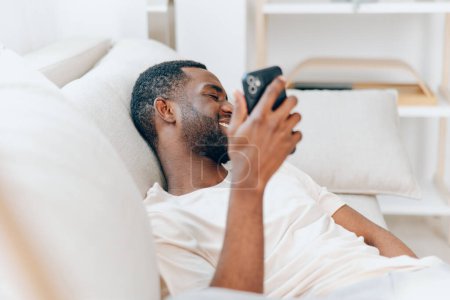 Foto de Feliz hombre afroamericano sentado en un sofá negro en su apartamento moderno, sosteniendo un teléfono inteligente y escribiendo un mensaje Está relajado y disfrutando de su tiempo libre, conectándose con amigos y navegando - Imagen libre de derechos