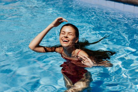 Téléchargez les photos : Femme heureuse nageant dans la piscine en maillot de bain rouge avec les cheveux longs lâches au soleil, protection de la peau avec écran solaire, concept de détente en vacances dans le climat tropical. Photo de haute qualité - en image libre de droit