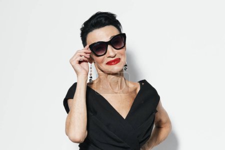 Foto de Mujer con estilo en vestido negro y gafas de sol posando con confianza con las manos en las caderas para la cámara de captura de aspecto de moda - Imagen libre de derechos