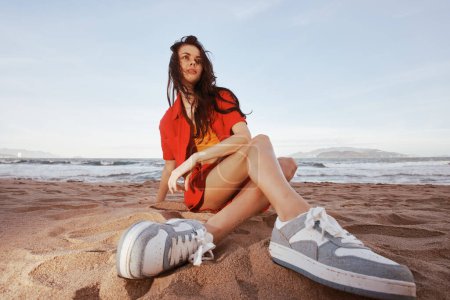 Téléchargez les photos : Femme insouciante souriante, assise sur la plage, profitant de vacances en tenue d'été tendance - Portrait de bonheur et de beauté au bord de l'océan - en image libre de droit