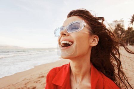 Téléchargez les photos : Évasion colorée au coucher du soleil : une femme souriante à la bouche ouverte, embrasser la liberté et la joie à la plage, profiter de vacances insouciantes dans des vêtements rouges à la mode - en image libre de droit