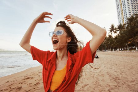 Téléchargez les photos : Vibrant Sunset Joy : Une femme souriante aux lunettes de soleil, embrasser la liberté et le bonheur, à la belle plage avec les vagues de l'océan. - en image libre de droit