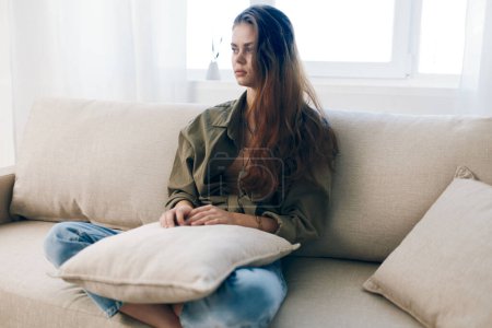 Téléchargez les photos : Femme inquiète assise sur un canapé, se sentant triste et stressée - Dépeignant des problèmes de santé mentale à la maison - en image libre de droit