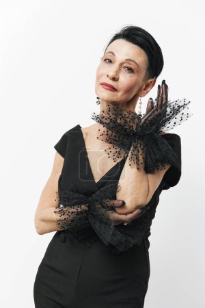 Elegant mature woman in black dress and gloves posing confidently for the camera with hands on hips
