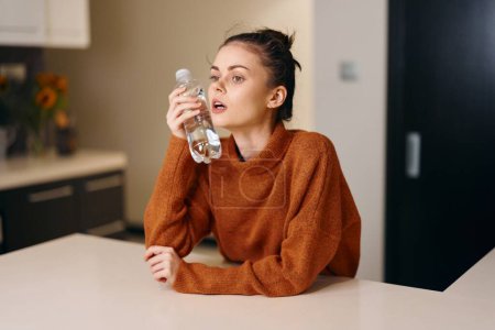 Foto de Enfermera sentada en casa sosteniendo un vaso de agua, retrato de una enferma hembra con un síntoma de resfriado y gripe, primer plano interior de una joven caucásica estornudando y bebiendo té de vitaminas - Imagen libre de derechos