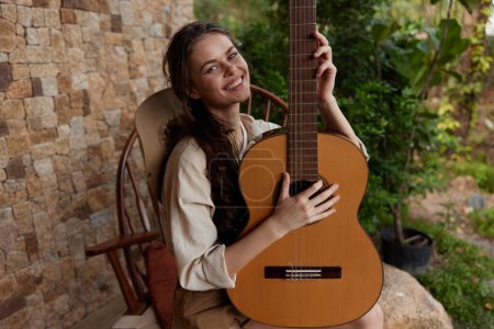 Téléchargez les photos : Une femme tient une guitare acoustique alors qu'elle est assise dans un fauteuil à bascule devant un mur de pierre - en image libre de droit