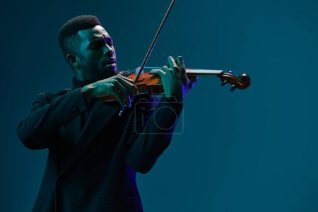 Téléchargez les photos : Violoniste professionnel en costume noir élégant jouant de la musique classique sur violon sur fond bleu - en image libre de droit