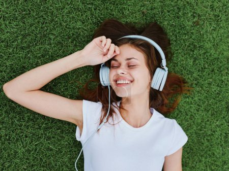 Téléchargez les photos : Une femme heureuse allongée sur l'herbe verte de la pelouse écoutant de la musique dans son casque souriant. Photo de haute qualité - en image libre de droit
