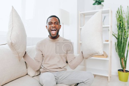 Téléchargez les photos : Homme afro-américain détendu assis sur un canapé confortable dans son appartement moderne, profitant d'un week-end paisible à la maison Avec un sourire sur son visage, il repose sa tête sur un oreiller, pensant et tenant un - en image libre de droit