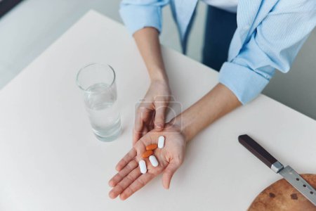 Photo for Woman holding pills next to a knife and glass of water on a table in a conceptual image about medication and health - Royalty Free Image