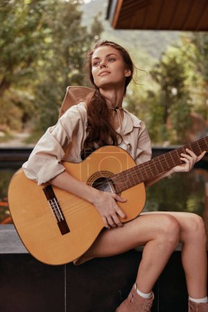 Photo for A young woman is sitting on a bench with an acoustic guitar in front of a pond - Royalty Free Image