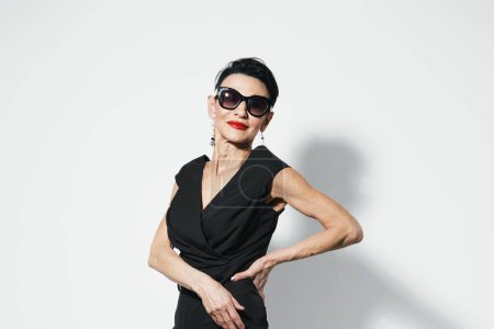 Stylish senior woman in black dress and sunglasses posing confidently with hands on hips for camera shot