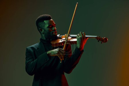 Photo for Elegant African American man in suit playing violin in front of dark background, creating beautiful music atmospher - Royalty Free Image