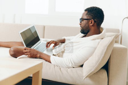 Téléchargez les photos : Souriant afro-américain indépendant en peignoir, travaillant sur un ordinateur portable sur le canapé dans sa maison moderne Profitant d'une matinée productive dans le cyberespace, tirant parti de la technologie pour son style de vie millénaire - en image libre de droit