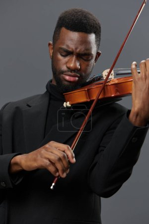 Téléchargez les photos : Afro-Américain en costume noir jouant du violon sur fond gris dans une performance musicale élégante et passionnée - en image libre de droit