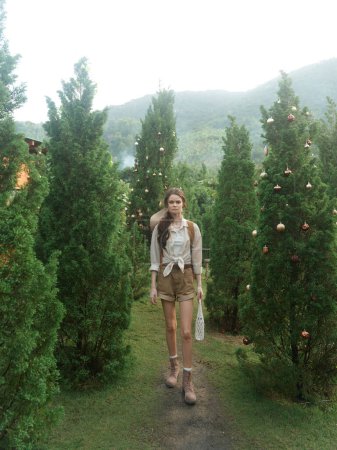 A woman in a white shirt and brown shorts walking through a forest of evergreen trees