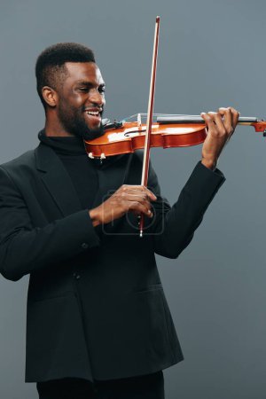 Téléchargez les photos : Musicien afro-américain en costume noir jouant du violon sur fond gris dans un concept de performance élégant - en image libre de droit