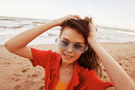Téléchargez les photos : Femme souriante dans les lunettes de soleil à la mode : Portrait d'une beauté insouciante Faire place à la liberté et à la joie à la plage colorée, avec le coucher de soleil magnifique sur l'océan comme toile de fond - en image libre de droit