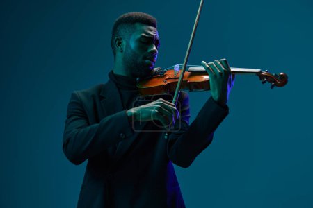 Photo for Elegant musician performing on violin in black suit against vibrant blue background - Royalty Free Image