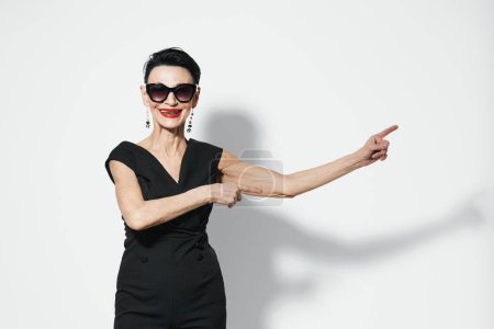 Téléchargez les photos : Femme élégante en robe noire et lunettes de soleil pointant du doigt quelque chose, isolée sur fond blanc - en image libre de droit