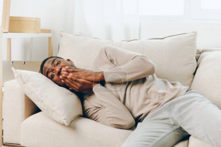 Foto de Hombre afroamericano estresado experimentando dolor de cabeza en un sofá en su casa Esta imagen captura la frustración y el dolor de un hombre joven mientras lidia con una migraña sentado solo en su sala de estar, él - Imagen libre de derechos