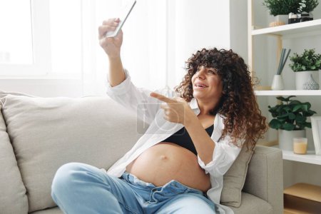 Foto de La bloguera embarazada se sienta en el sofá de su casa y se toma fotos por teléfono, selfies y videollamadas, consulta con el médico en línea, manejo del embarazo. Foto de alta calidad - Imagen libre de derechos