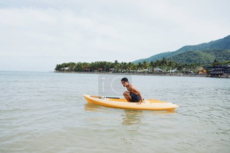 Photo for Happy Asian man enjoying kayaking in azure tropical waters with palm-fringed beach as backdrop. - Royalty Free Image