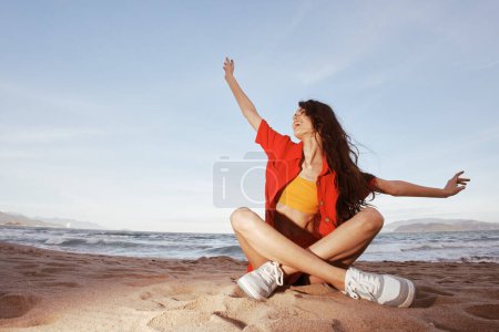 Téléchargez les photos : Femme insouciante assise sur une plage de sable, profitant de vacances d'été à la mode avec une vue grand angle sur le bel océan - en image libre de droit
