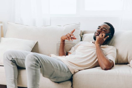 Foto de Hombre afroamericano feliz hablando por teléfono mientras está sentado en el sofá negro en el apartamento moderno - Imagen libre de derechos