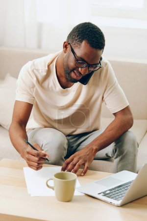 Photo for Young African American man sitting on a comfortable sofa in his modern apartment, working on his laptop with a happy and focused expression He is a freelance worker, using cyberspace and modern - Royalty Free Image