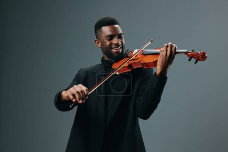 Téléchargez les photos : Elégant jeune homme afro-américain en costume noir jouant du violon sur fond gris au studio - en image libre de droit