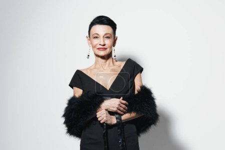 Elegant older woman in black dress and fur stole posing for a photo in front of white wall