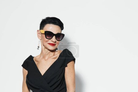 Photo for Confident woman in elegant black dress and sunglasses posing with hands on hips in front of white wall - Royalty Free Image