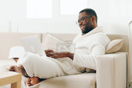 Téléchargez les photos : Smiling African American Freelancer Dactylographier sur ordinateur portable dans le bureau moderne, profiter du travail du matin Le jeune homme noir est habillé d'un peignoir confortable, assis sur un canapé, et de travailler avec diligence sur - en image libre de droit