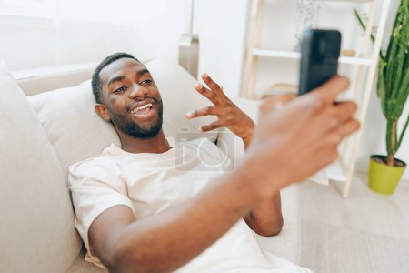 Téléchargez les photos : Happy African American Man assis sur un canapé noir et utilisant un téléphone portable dans son appartement moderne Il est détendu et souriant tout en tapant un message, profitant du confort et de la connexion fournis par - en image libre de droit