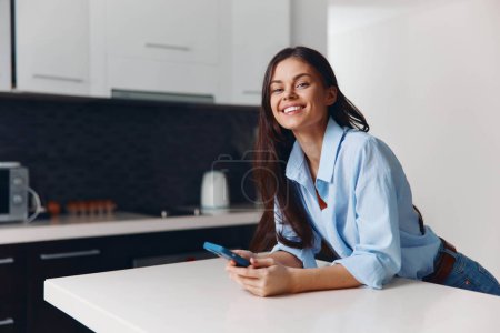 Foto de Mujer feliz sentada en el mostrador de la cocina charlando en el teléfono celular con una sonrisa en su cara - Imagen libre de derechos