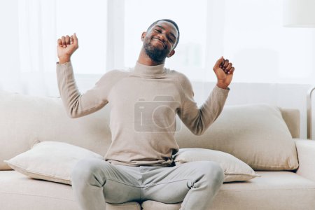Téléchargez les photos : Homme afro-américain souriant assis sur le canapé, profitant d'un week-end relaxant à la maison Il se repose confortablement sur le canapé, tenant un oreiller et pensant avec un regard de contentement La vie moderne - en image libre de droit
