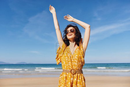 Téléchargez les photos : Femme joyeuse dans une robe jaune ensoleillée célébrant sur une belle plage de sable avec les bras levés haut dans les airs - en image libre de droit