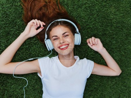 Foto de Retrato de mujer tumbada en la hierba verde en un soleado día de verano escuchando música en auriculares con una camiseta blanca, sonrisa con dientes felicidad mirar a la cámara. Foto de alta calidad - Imagen libre de derechos