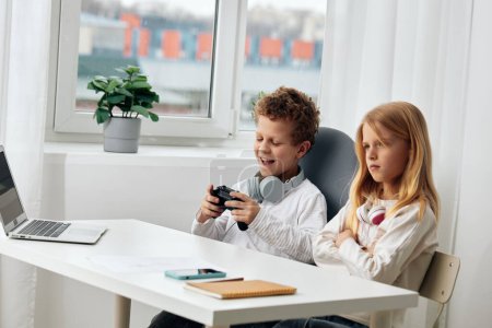 Photo for Happy caucasian boy and girl sitting at a table in their living room, engrossed in their online elearning session The siblings are wearing headphones and using their laptops and phones to connect to - Royalty Free Image
