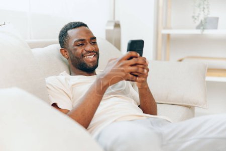 Téléchargez les photos : Heureux homme afro-américain se détendre sur le canapé noir, Dactylographier un message sur un téléphone portable dans un appartement moderne bénéficiant de la technologie et de la connexion, souriant et se reposant à la maison - en image libre de droit