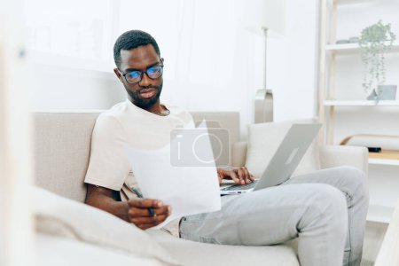 Photo for Smiling African American Man Working on Laptop in Modern Home Office, Typing and Chatting Online This image captures a young and happy freelancer, sitting on a sofa in his cozy living room He is an - Royalty Free Image