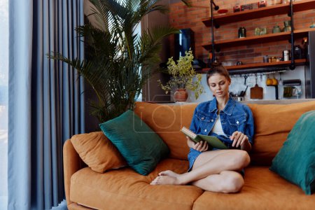 Foto de Mujer relajante en el sofá naranja cerca de libro de lectura de la ventana en acogedora sala de estar con luz del día brillando a través de - Imagen libre de derechos