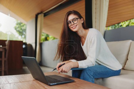 Téléchargez les photos : Femme souriante assise sur un canapé avec ordinateur portable au bureau à domicile Concept d'éducation et de travail en ligne - en image libre de droit