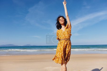Foto de Mujer en vestido amarillo con los brazos levantados de pie en la playa disfrutando del sol y la libertad - Imagen libre de derechos