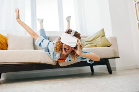 Foto de Mujer sonriente disfrutando de un juego de realidad virtual en casa con gafas VR futuristas en el sofá - Imagen libre de derechos