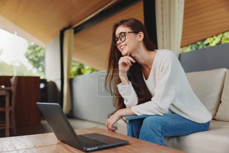 Photo for Smiling Woman Using Laptop on Sofa in Home Office A young woman sits comfortably on a sofa in her living room, wearing glasses and a smile on her face She is a teacher, conducting an online class - Royalty Free Image
