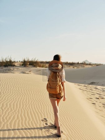 Woman hiking through sunlit desert sand dunes with backpack on a clear sunny day